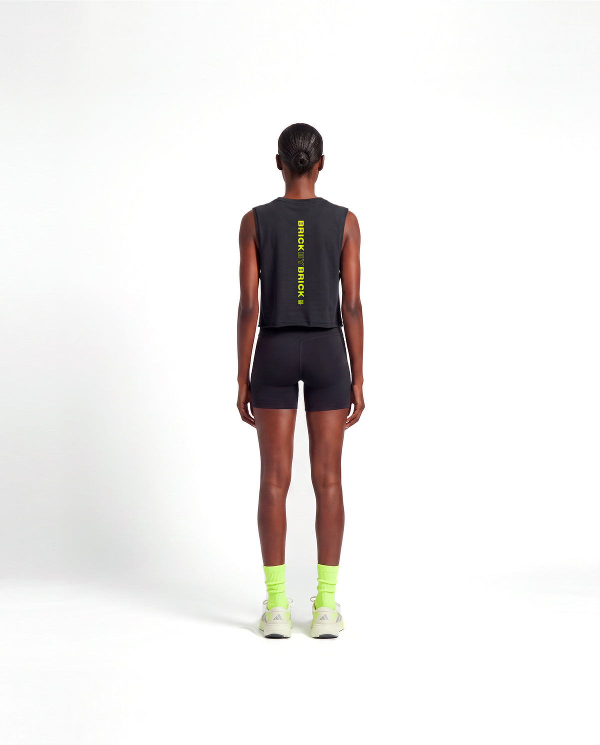 Model wearing the Brick by Brick cropped tank top in black — back view with BRICK BY BRICK set vertically down the centre in lime green — View 4