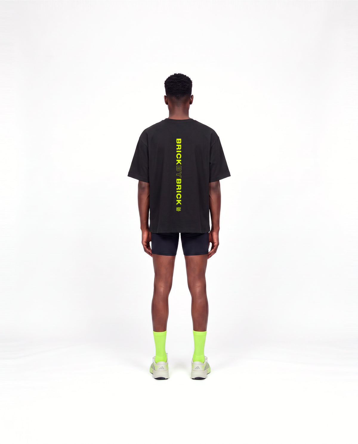 Model wearing the Brick by Brick oversize t-shirt in black — back view with BRICK BY BRICK set vertically down the centre in lime green — View 4