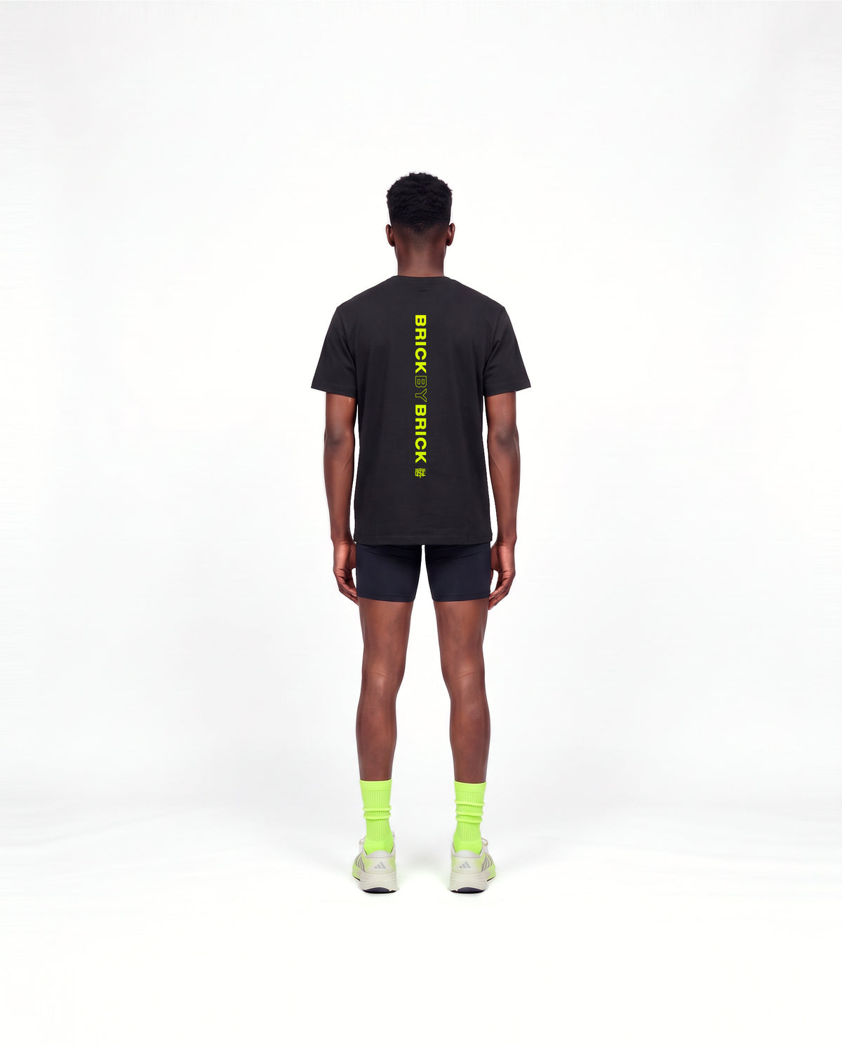 Model wearing the Brick by Brick t-shirt in black — back view with BRICK BY BRICK set vertically down the centre in lime green — View 4
