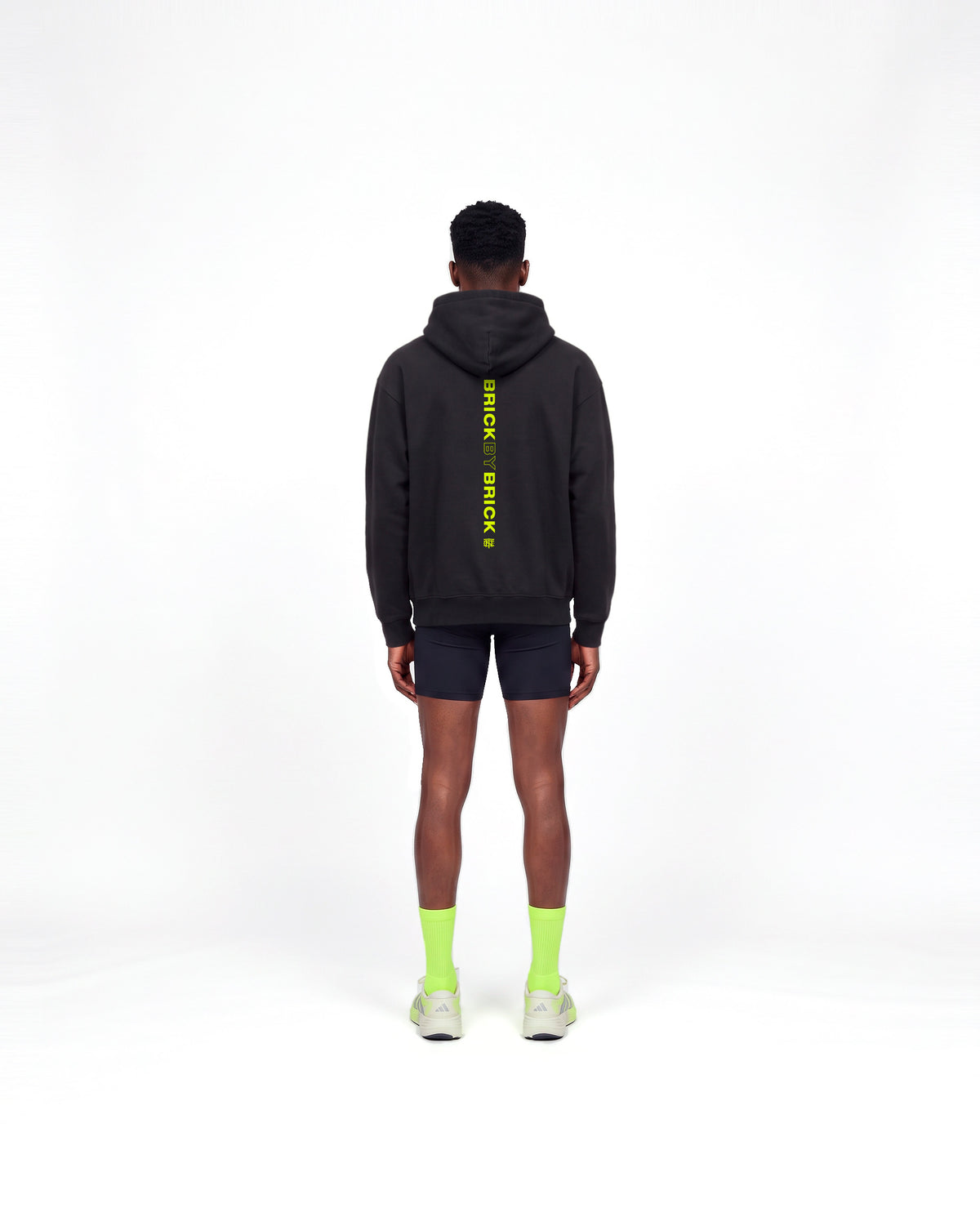 Model wearing the Brick by Brick oversize hoodie in black — back view with BRICK BY BRICK set vertically down the centre in lime green — View 4
