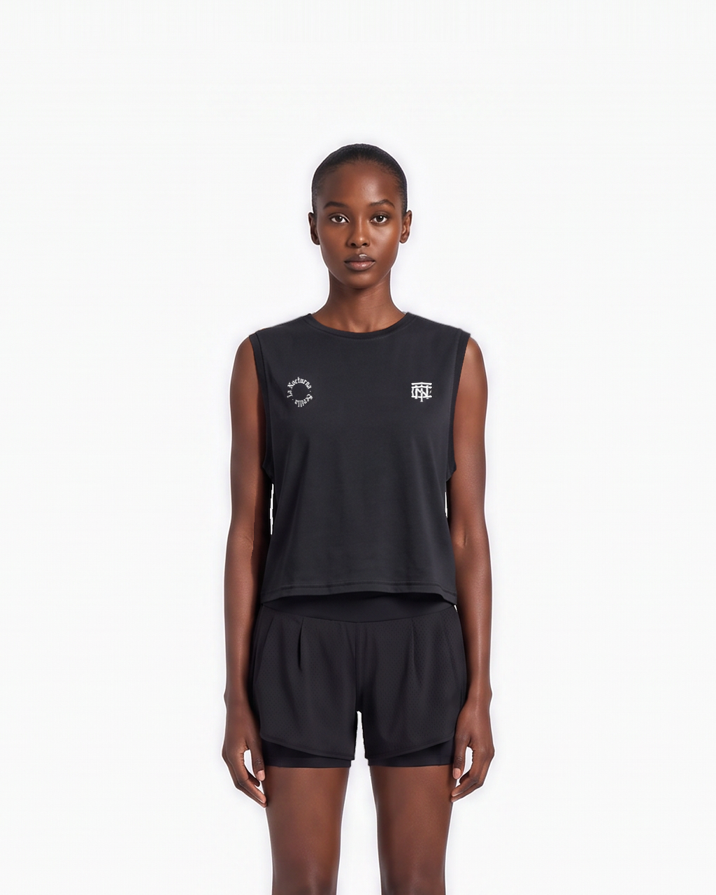 Model wearing the Let's Run the Night cropped tank top in black — front view, close crop on the La Nocturna Sevilla seal and the TN monogram in white — View 3
