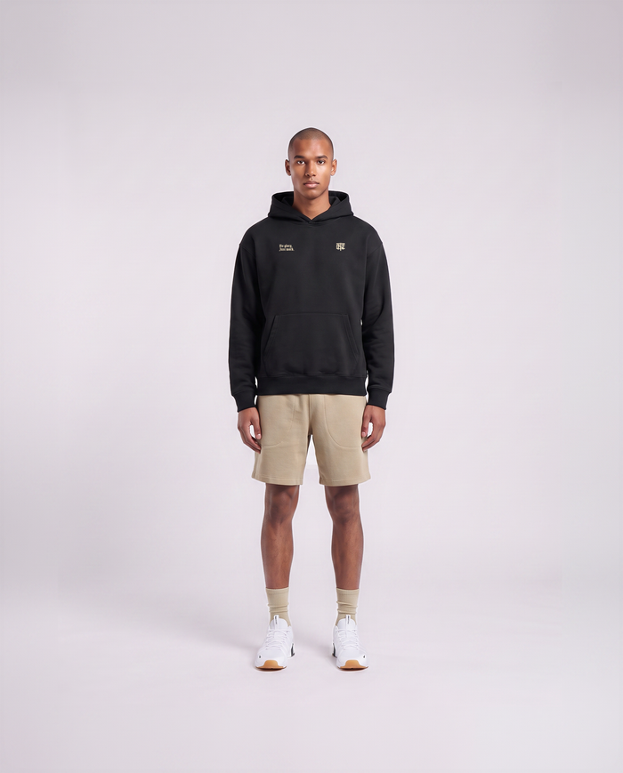 Model wearing the No Glory Just Work oversize hoodie in black — front view with the left-chest NO GLORY JUST WORK wordmark and the right-chest Thundernoise monogram in sand