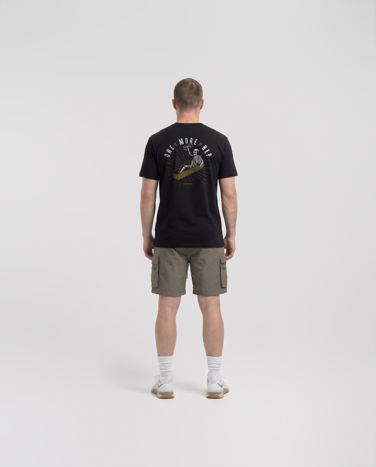Model wearing the One More Rep t-shirt in black — back view with the skeleton pressing one last dumbbell out of an open coffin under ONE MORE REP in bone white and gold — View 4

